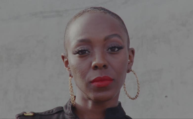 Bald female model with hoop earrings and short black dress faces the camera with eye contact