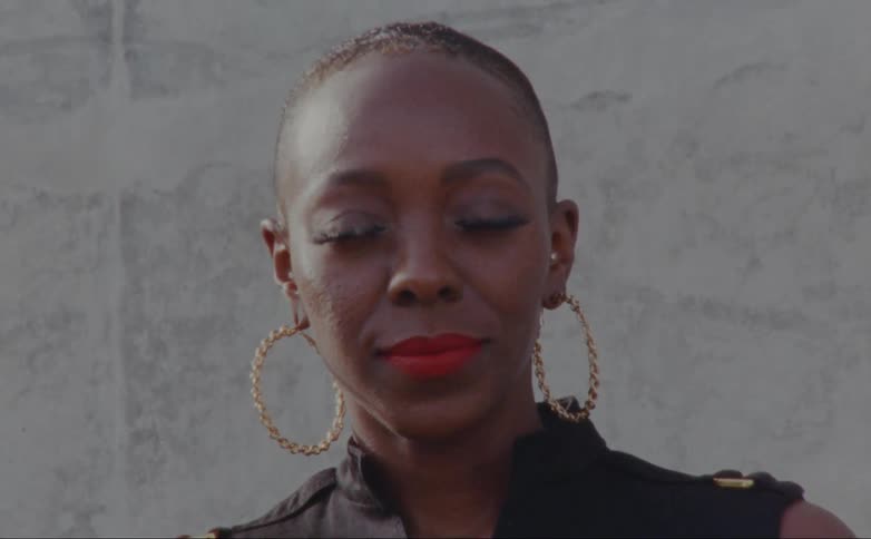 A black woman with large hoop earrings stands in front of a concrete wall