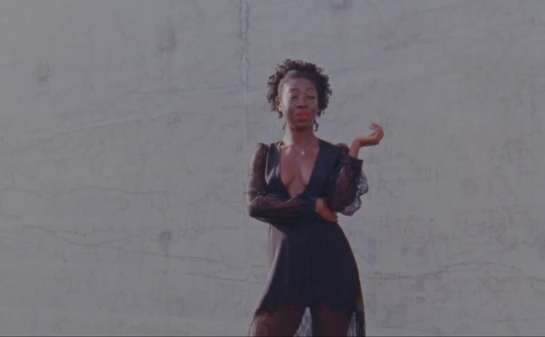 Female model with curly hair and black decollete dress poses for the camera in front of wall