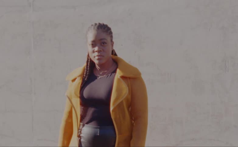 Woman with dreadlocks hoop earrings yellow overcoat and black shirt poses for the camera