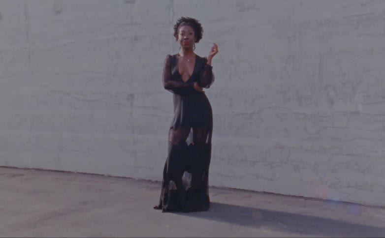 Female model with curly hair and black plunging gown poses for the camera in front of a wall outdoors