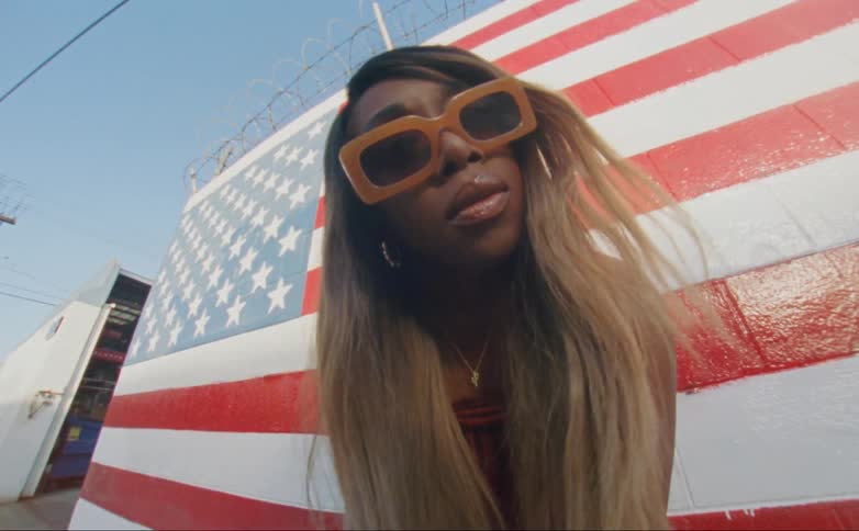 Black woman standing in front of a american flag painted wall wearing orange sunglasses during day time