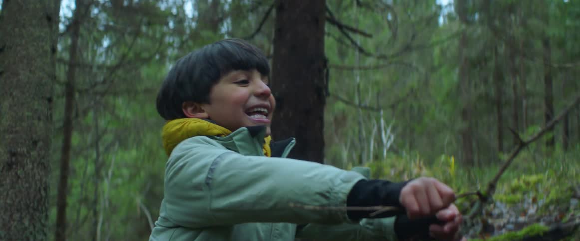 A young boy wearing a jacket is playing in a forest and holding a stick