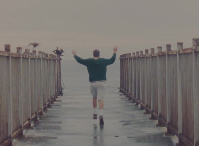 Man runs down walkway between wooden fencing toward the sea as seabird take off
