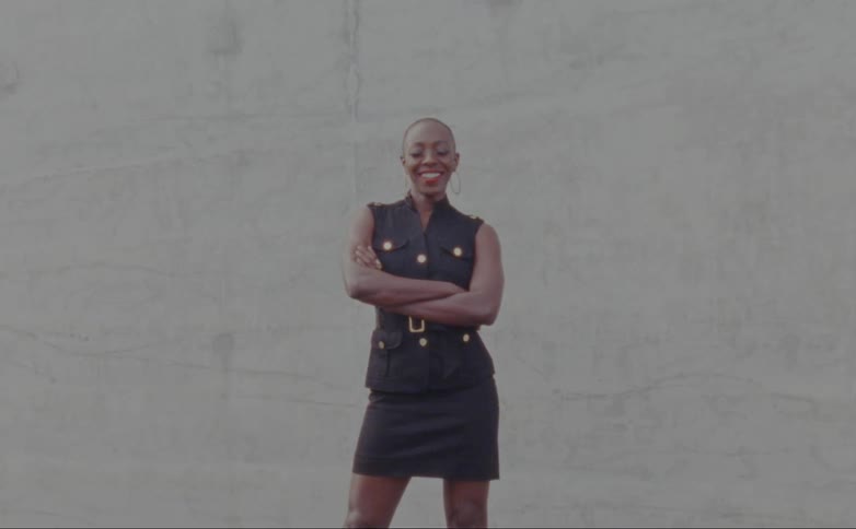 Woman with hoop earrings and black dress stands arms crossed in front of wall