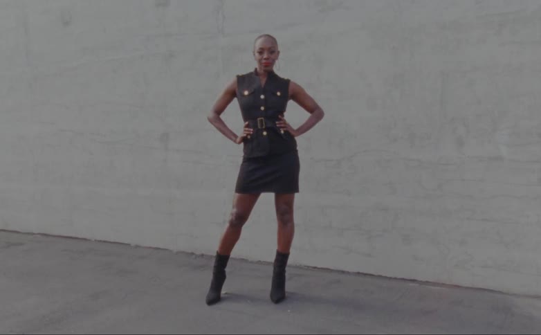 Woman with hoop earrings black short dress and boots poses for camera in front of wall
