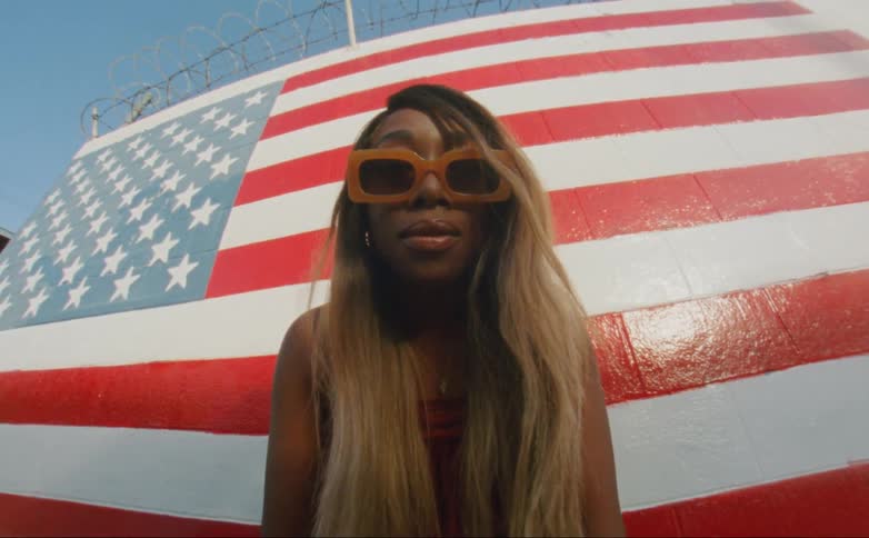 Black woman with long hair and wearing orange sunglasses stands in front of a large american flag