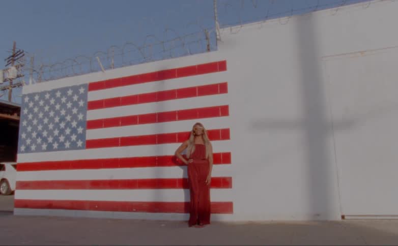 A woman wearing a long gown is posing in the evening in front of an american flag painted on the side of a building
