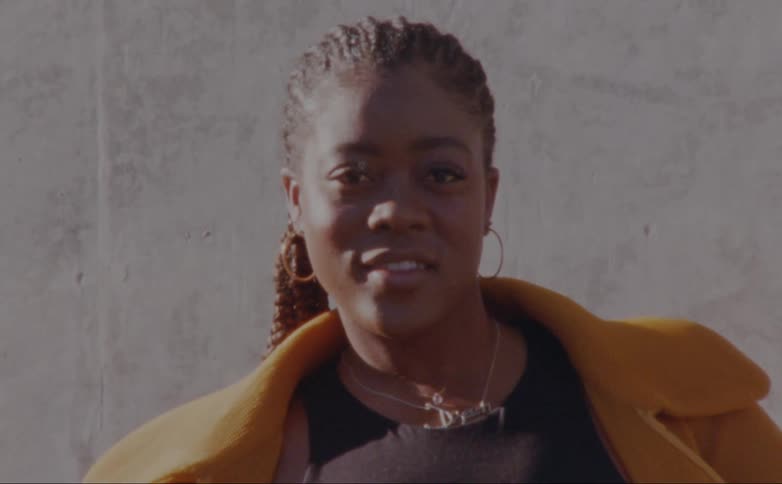 Woman with dreadlocks hoop earrings and yellow overcoat faces the camera with eye contact