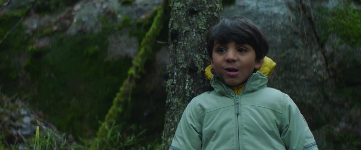 A young boy wearing a jacket is standing in a forest and he smiles as he puts something in his mouth and takes a bite