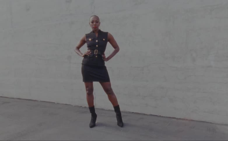 Black woman wearing black leather clothing and black boots posing for the camera in front of a grey wall