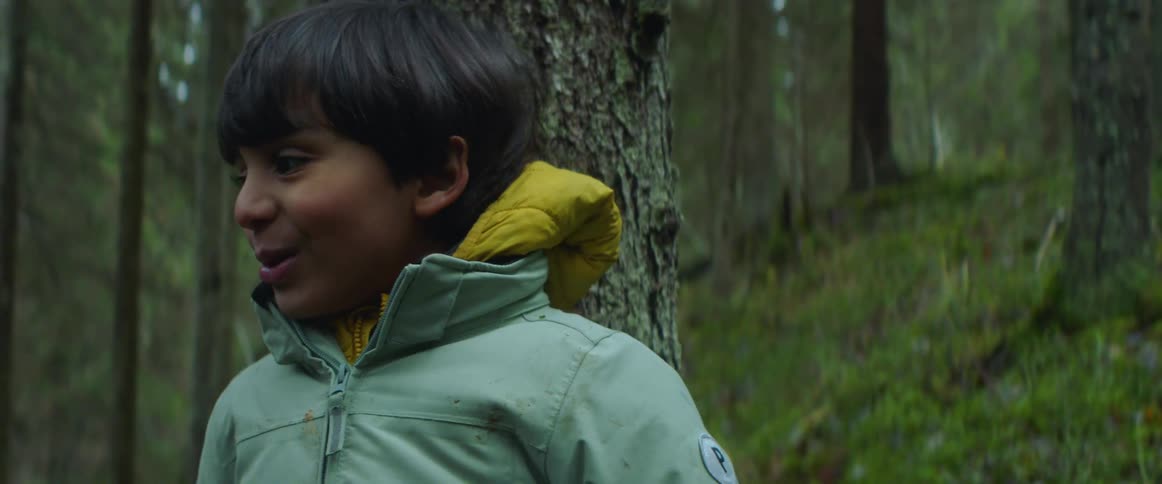 Young boy in a blue jacket smiles while standing in front of a tree in the forest