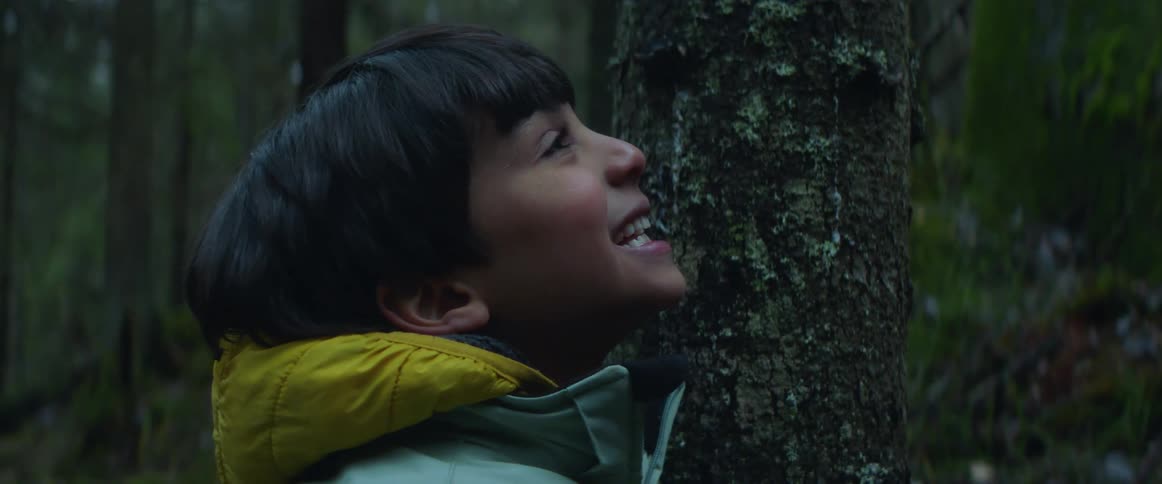 Young boy smiling and shaking his head while standing at the middle of a forest next to a tree trunk wearing a coat