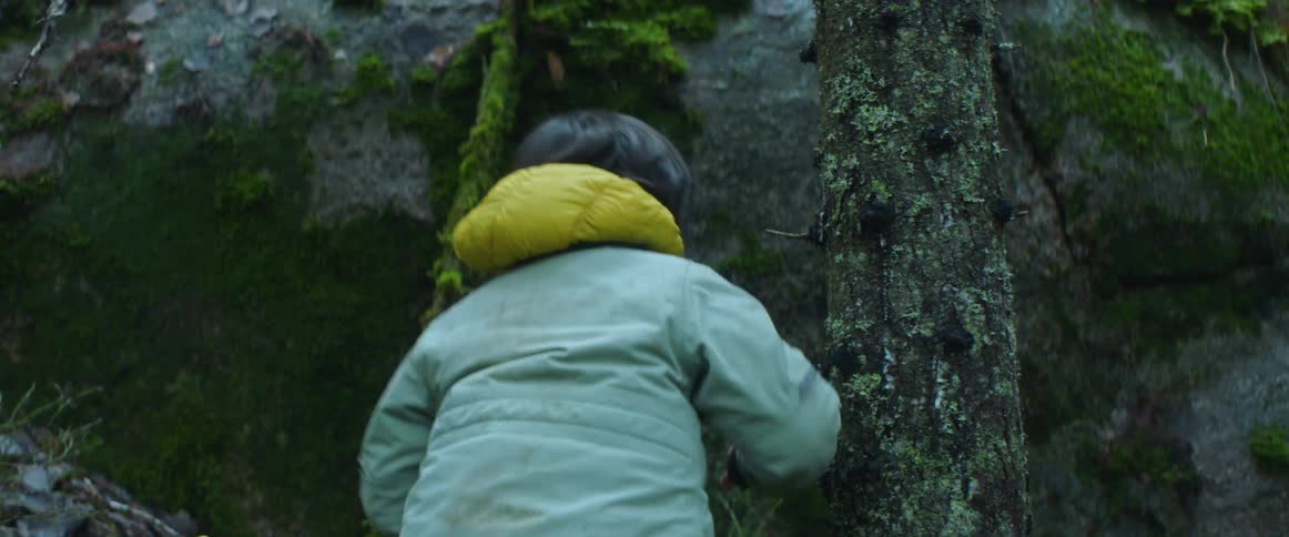 Mother and son in the forest as the boy carries a stick towards the large rock and turns around