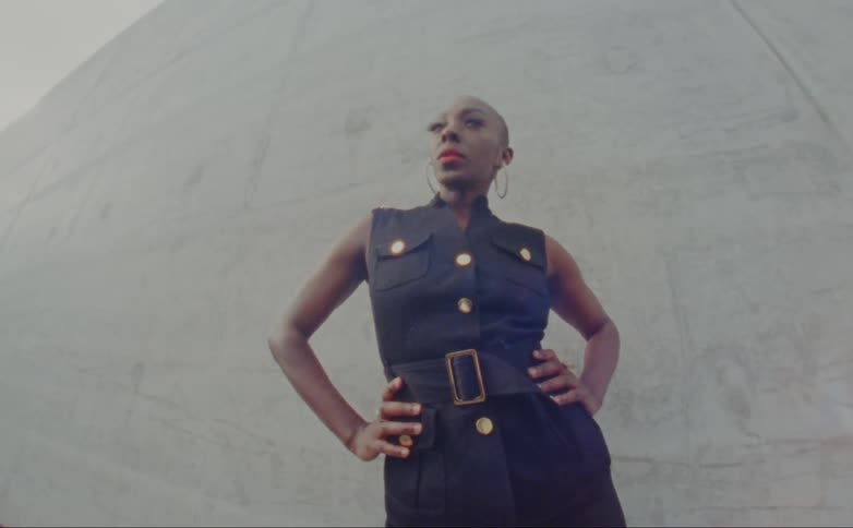 Bald female model with hoop earrings and black dress stands in front of wall