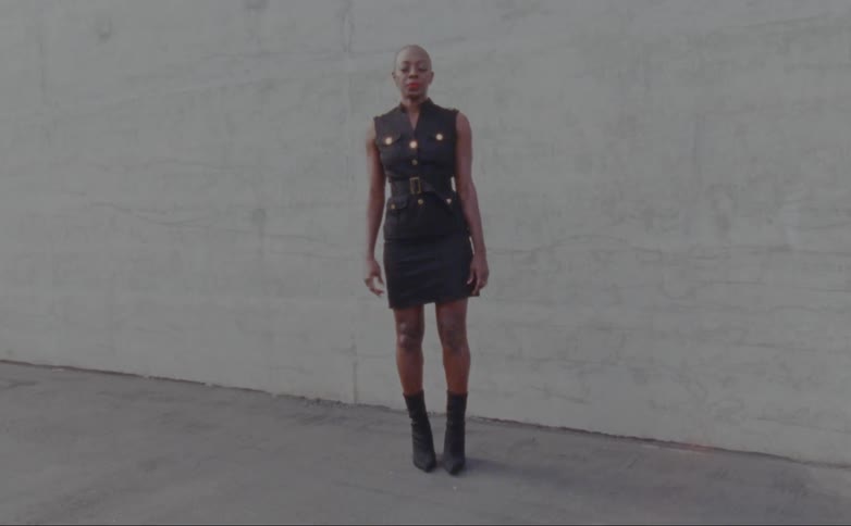 Woman with hoop earrings black short dress and boots poses for camera in front of wall