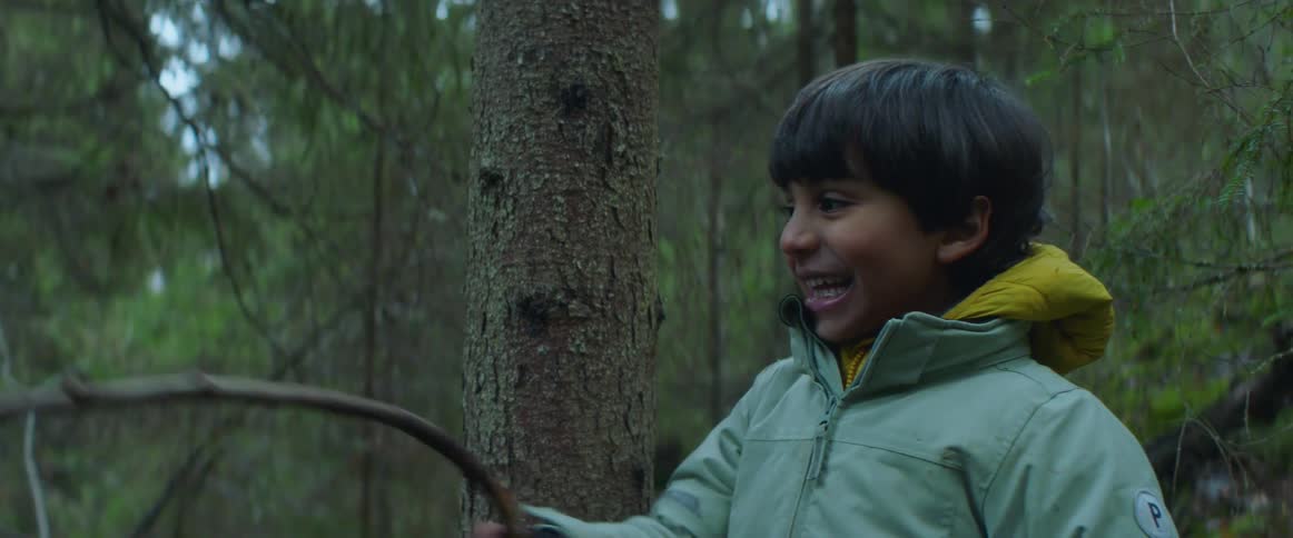 A young boy is playing in the forest near a tree and waving a stick
