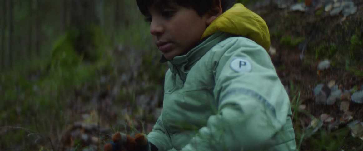 Boy with turquois jacket and jeans sits on slope in the woods