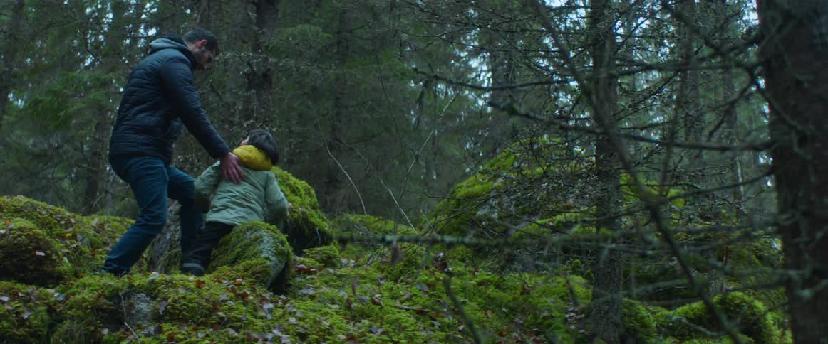 Father and young son wearing jackets climb over the mossy rocks in the forest