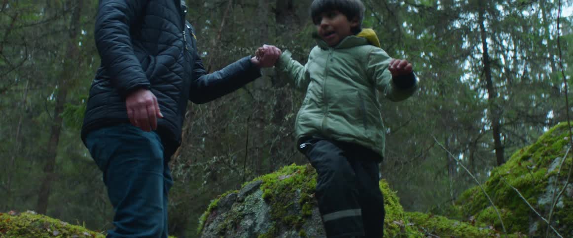 Father holds his sons hand as the son jumps off of a mossy rock in the forest