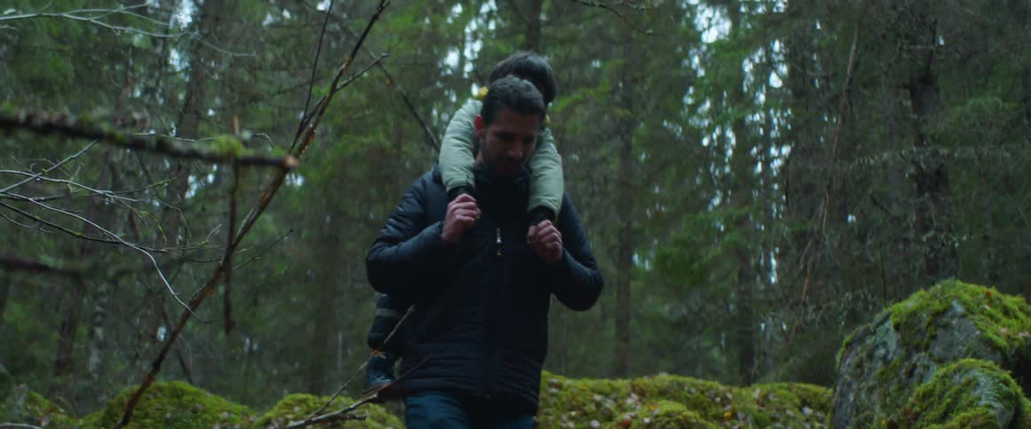 Man carries a little boy on his back as he climbs down the mossy rocks in the forest