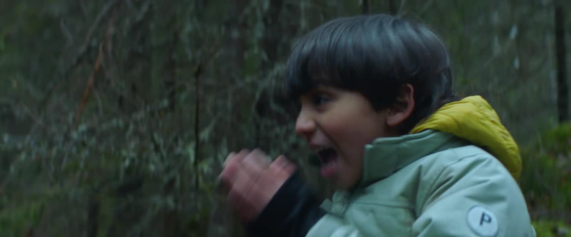 A young boy wearing a jacket is standing in a forest as he says something and covers his mouth with his hands