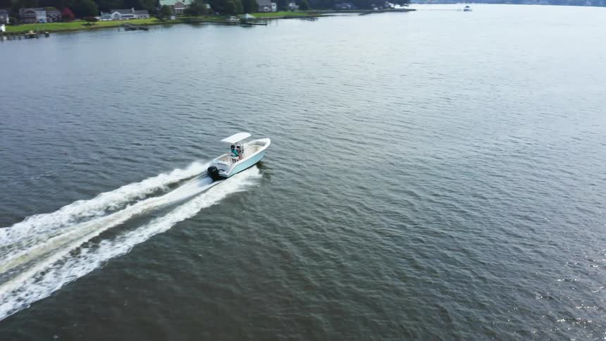 People pilot boat through water paralleling shore and passing homes