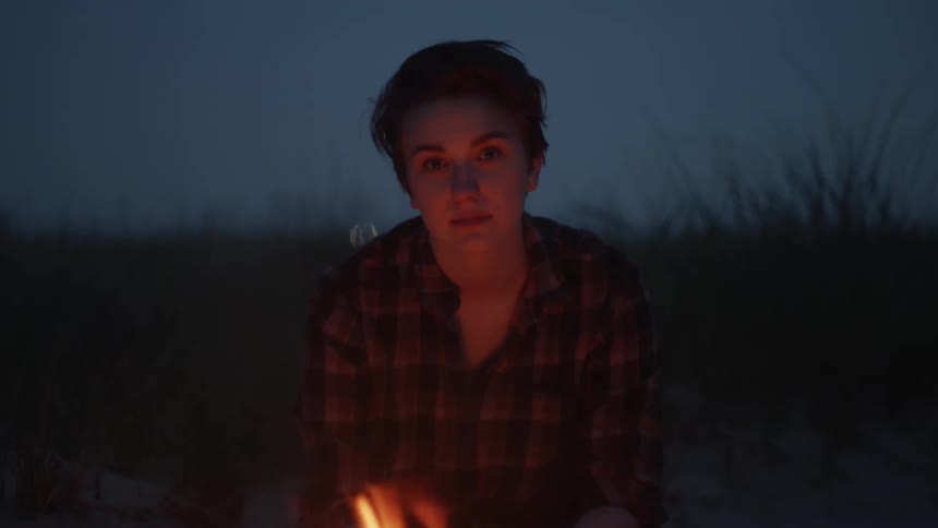 Girl with short hair and plaid shirt sits near bonfire looking at camera at night