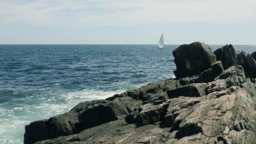 Sailboat navigating at the sea in a sunny day while tides are hitting the coastal rocks