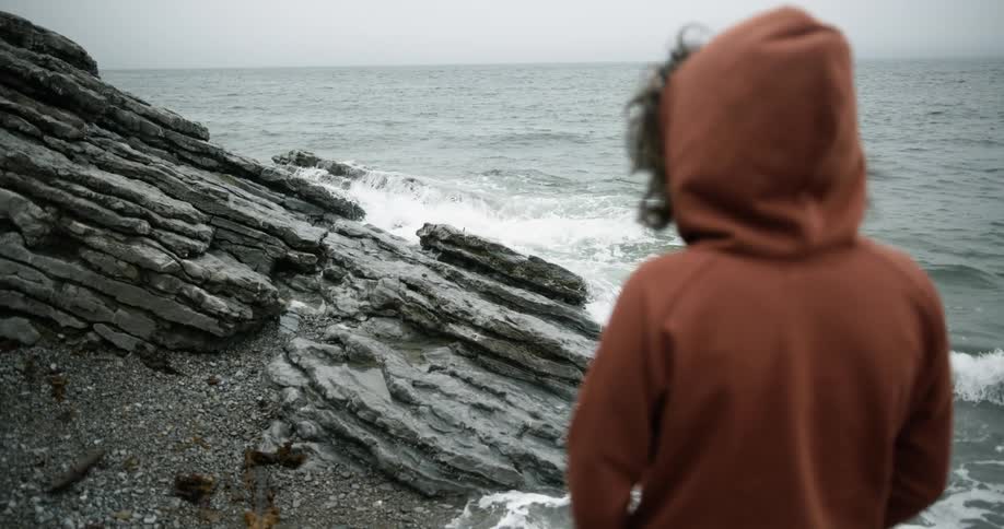 A woman wearing a hooded jacket is standing along the coast watching the waves crash into the rocks as they roll in