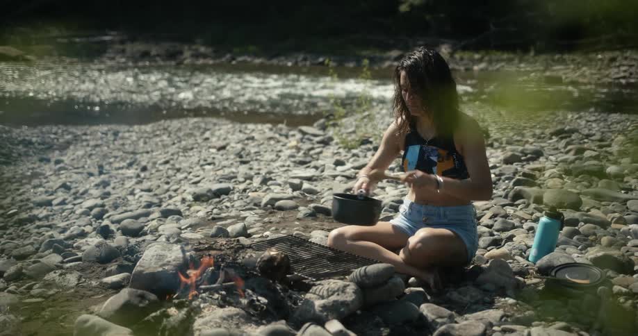 A woman sits on a rocky beach cooking over a campfire beside a river