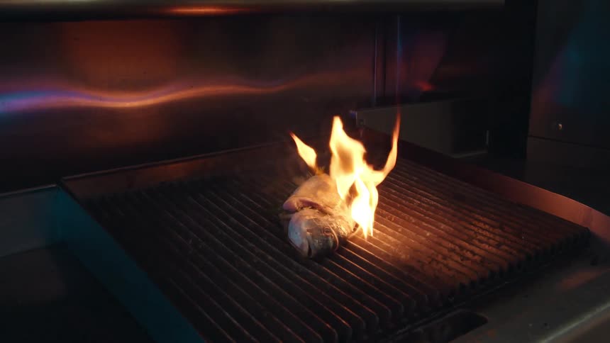 Flames curve over fish lying on the grill in a professional metallic kitchen