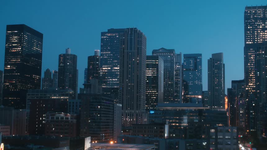 A city skyline with skyscraper windows lit at dusk and headlights glowing from the street