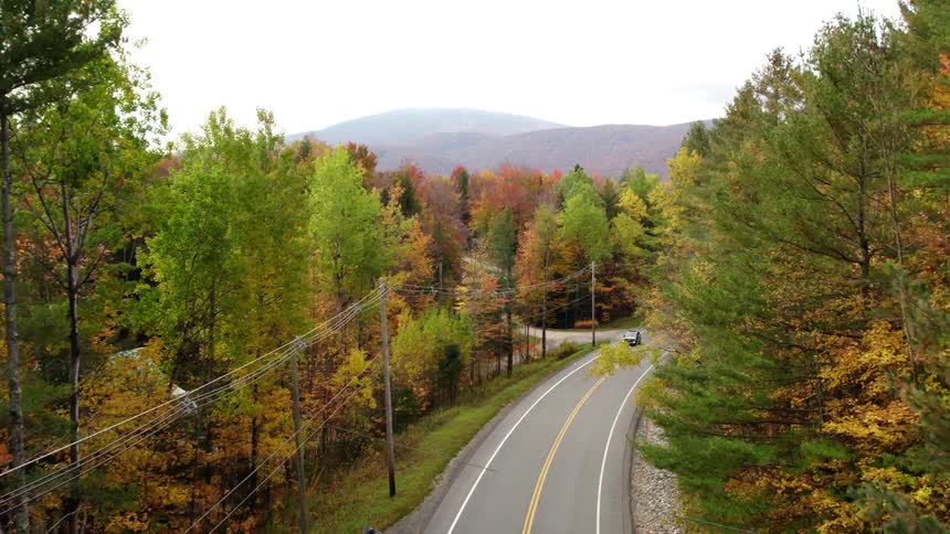 An automobile travels on a road through forest of trees with bright fall colored leaves and mountains in the distance