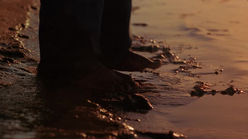 Close up mans bare feet wearing pants stepping onto shoreline at the lake