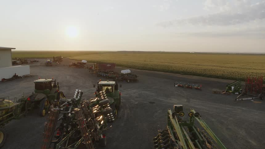Tractors and other farm equipment are parked near a vast field of crops as the warm morning sun hangs low in the sky