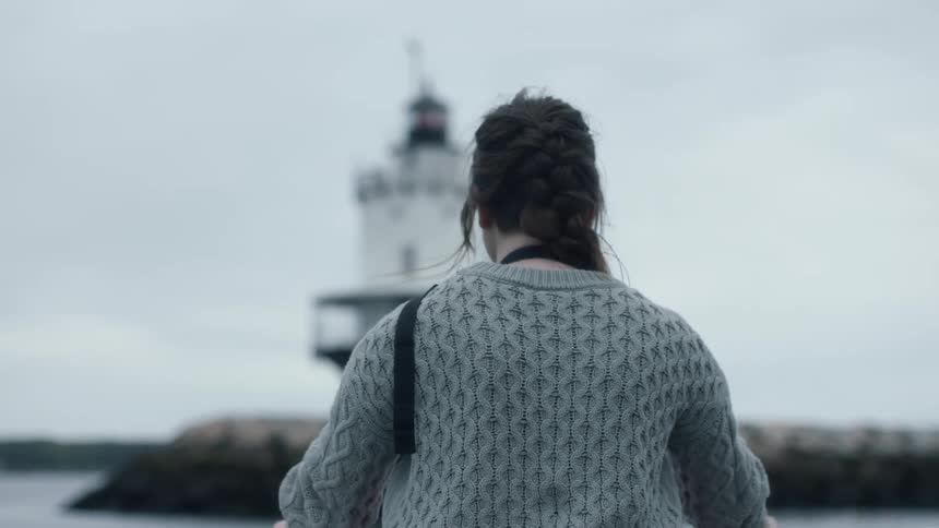 Woman stands on vessel as it bobs past lighthouse standing on nearby shore