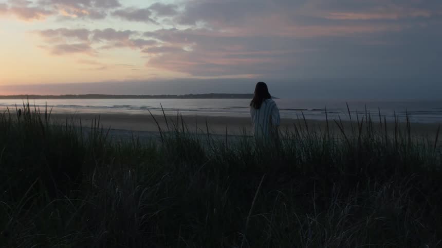 A girl walks through tall grass and weeds towards a beach at dusk