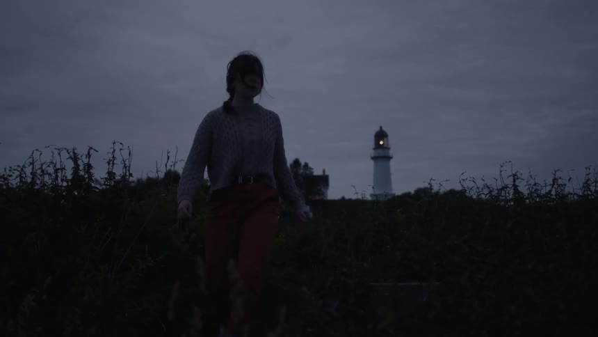 A girl is walking through the weeds along the coast as the beacon on a lighthouse shines