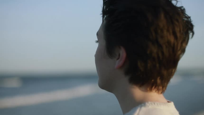 Woman walks towards the ocean then dips her hands in the water and runs her hands through her hair