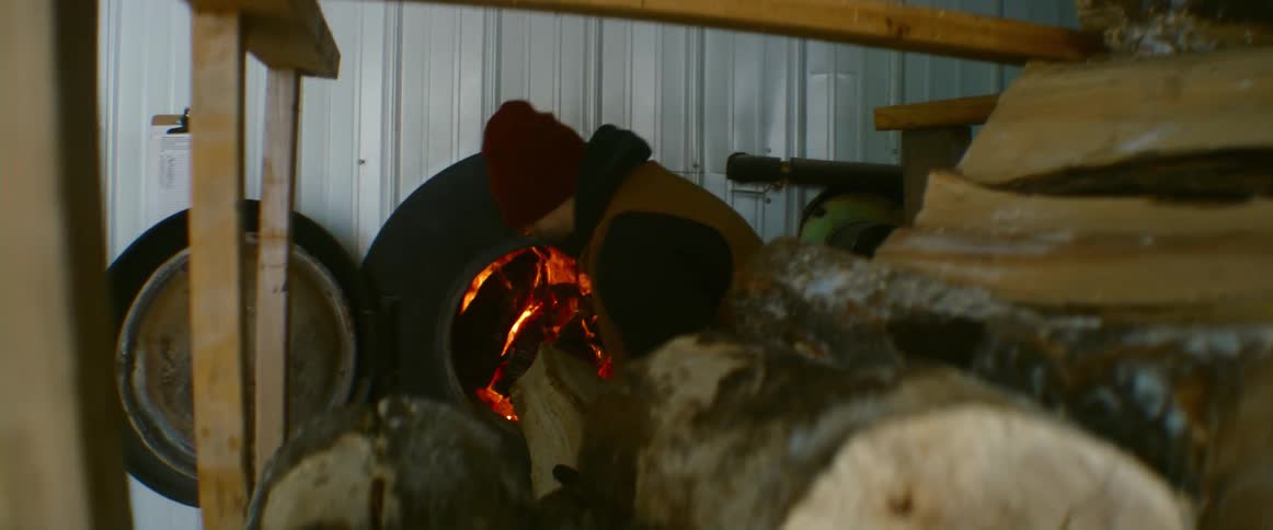 Man puts log from stack of ice coated firewood into furnace then closes door