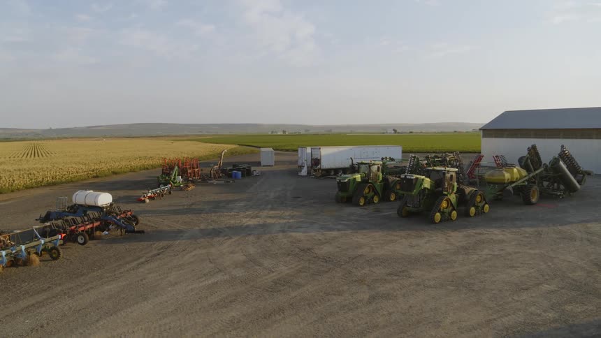 Farm machinery sits parked on a gravel area as multiple crop fields stretch to the mountains along the horizon