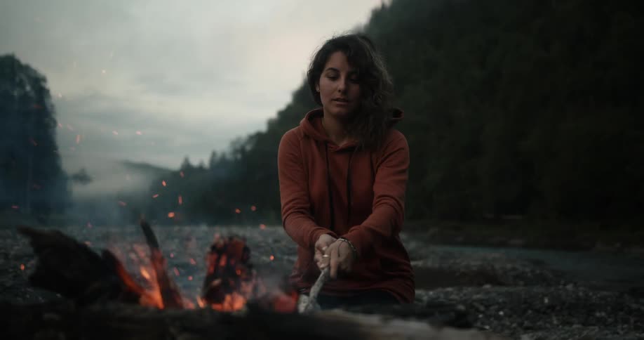 A woman by a forest stirs a campfire