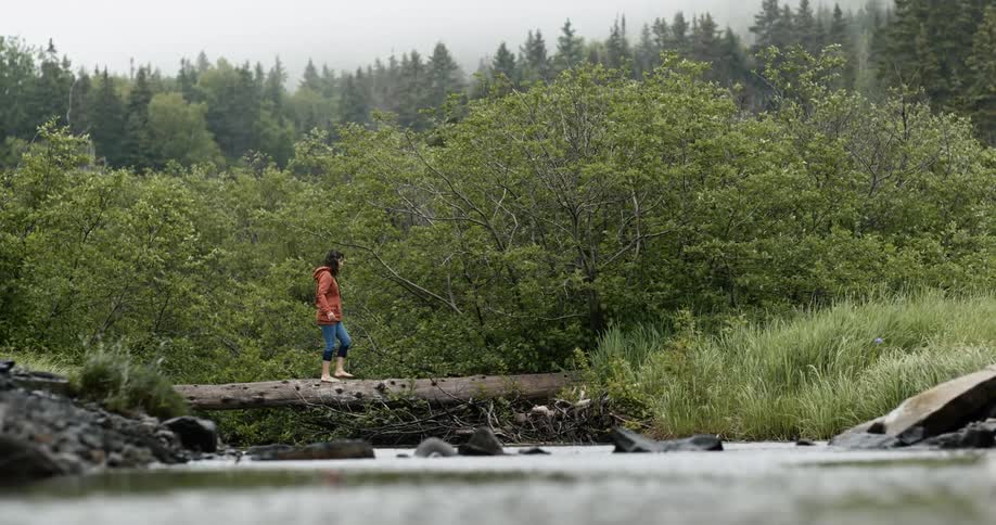 A woman crosses a river on a log bridge ducking under tree branches on the other side