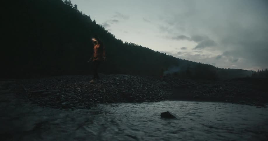 A woman is walking away from a campsite in the forest at dusk toward a stream and squats to get some water