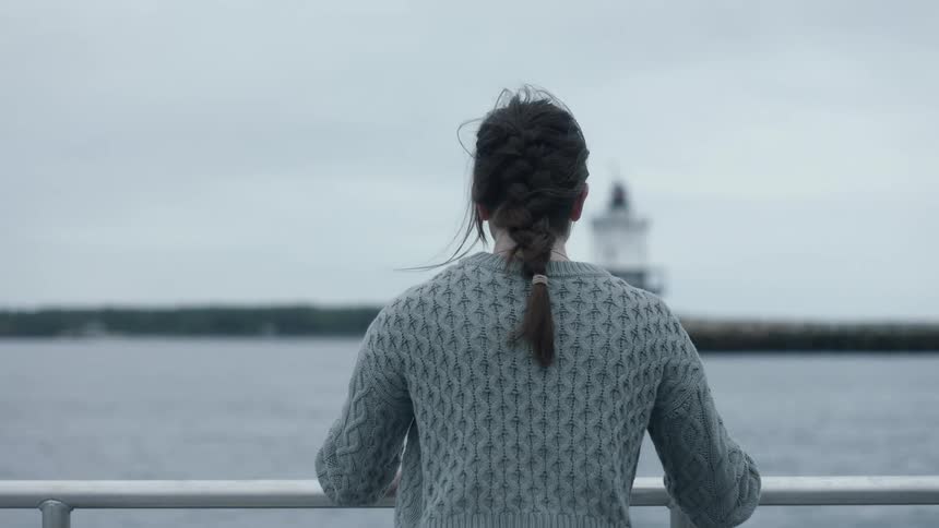 A woman traveling on a ship approaches a railing and looks out toward the shoreline on a windy and overcast day