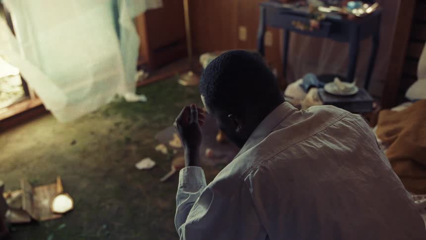 A fully dressed man sits up in bed rubbing his head in a dirty messy room