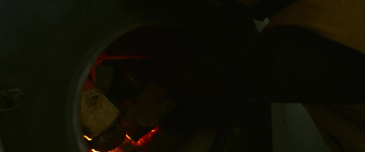 Man wearing a toque and a mask pulls firewood from a pile and puts it into the fire burning in a furnace