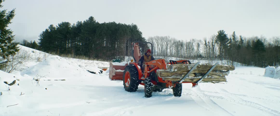A man on tractor carries logs on a front end loader in the snow