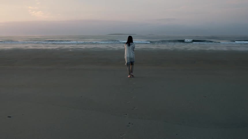 Woman wearing cardigan walking towards shoreline at the beach at sunrise